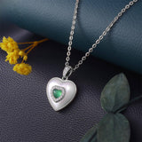 Silver heart-shaped necklace with a green gemstone on a dark textured surface with yellow flowers and green leaves.