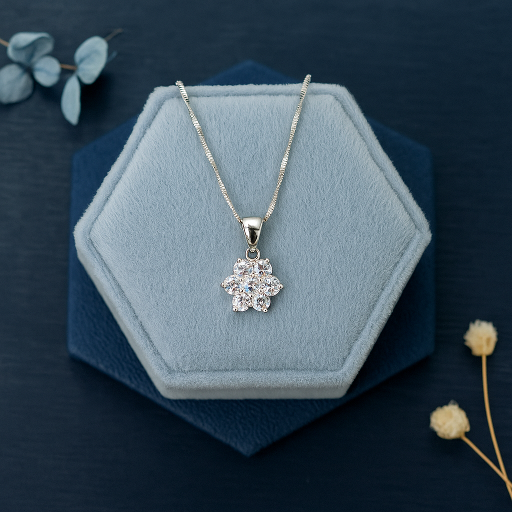 Silver necklace with a floral pendant on a blue hexagonal stand with flowers around.