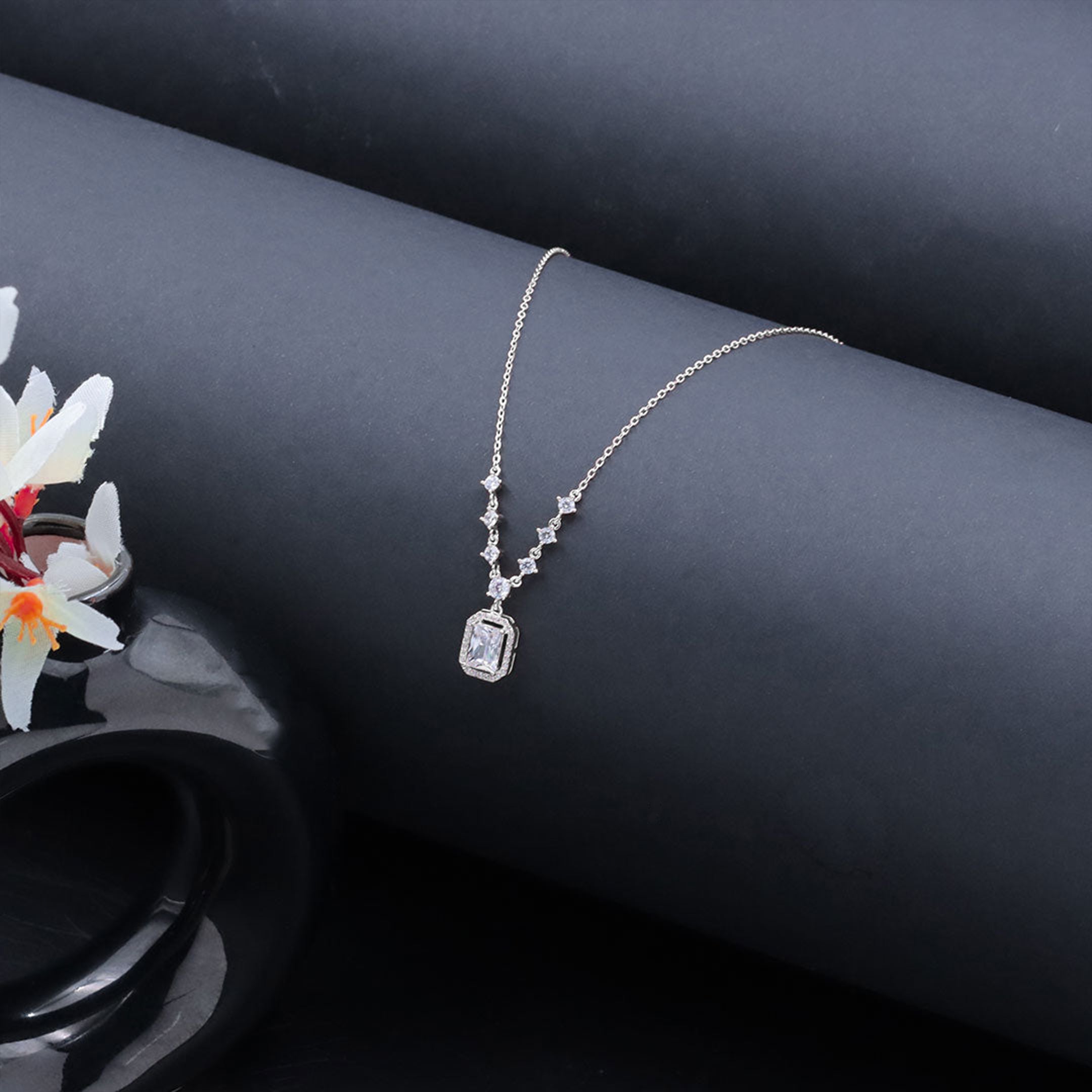 Silver necklace with a diamond pendant on a dark surface with flowers.
