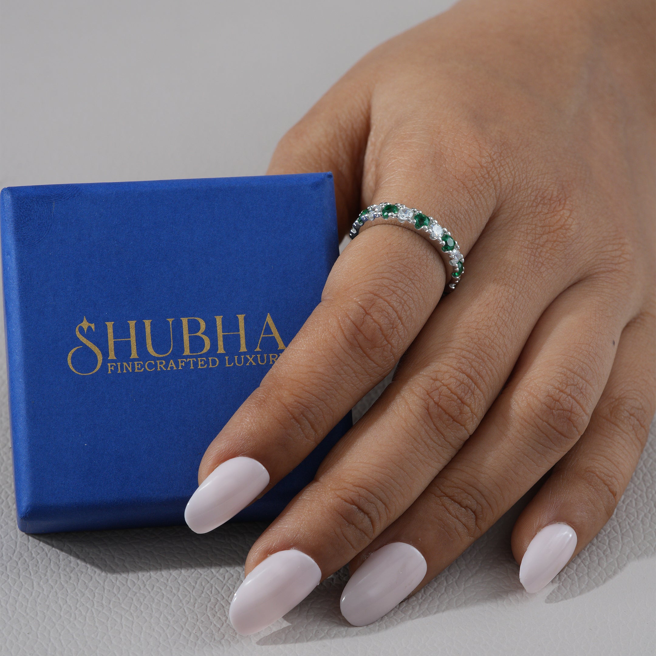 Hand wearing a ring with gemstones next to a blue Shubha box