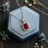 Silver necklace with a red gemstone pendant on a blue cushion against a dark wooden background.